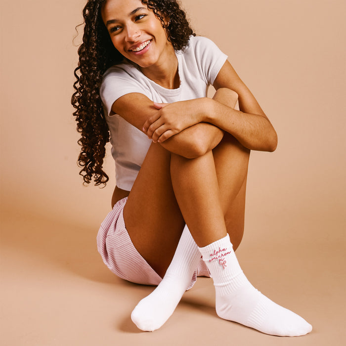 A smiling young woman with curly hair sits on the floor wearing a white T-shirt, pink striped shorts, and The Darling Effect Alpha Omicron Pi Embroidered Crew Socks with red writing, against a beige background.