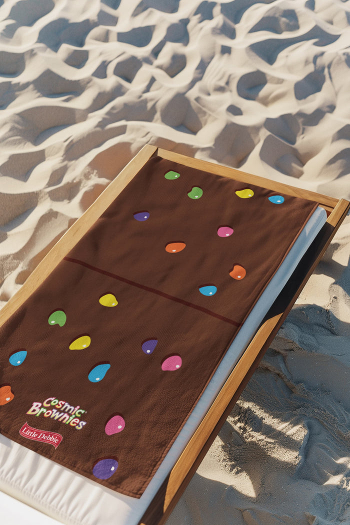A Little Debbie® Cosmic Brownie beach towel laid out flat on a wooden lounge chair over white cushions on a sandy beach. The towel resembles a Cosmic® Brownie with a chocolate-brown color and colorful, candy-like sprinkle shapes in various colors scattered across. The "Cosmic® Brownies" logo and a red Little Debbie® logo are positioned near the bottom of the towel. The sand around the chair is textured with shadows, showing the sunlit beach setting.