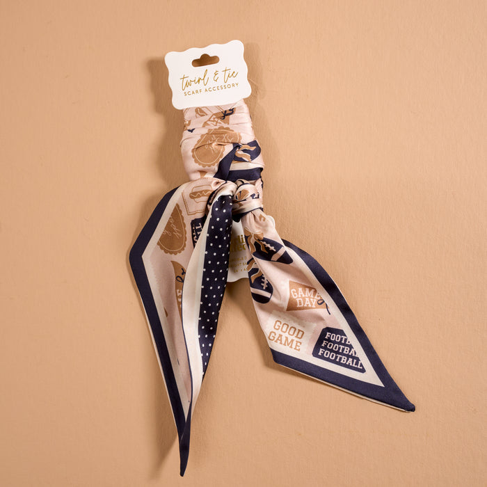 Introducing the Huddle Up Twirl & Tie Scarf Accessory by The Darling Effect—a silky satin scarf adorned with football-themed prints and bold phrases like "GOOD GAME" and "FOOTBALL FOOTBALL FOOTBALL" set against a beige backdrop. Presented on a branded white card, this accessory is perfect for adding sporty flair to your look.