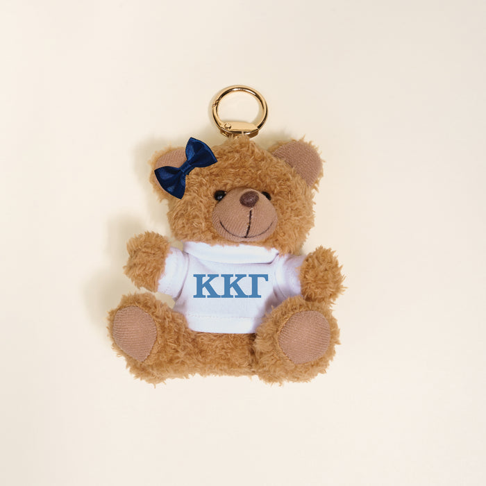 The Darling Effect's Kappa Kappa Gamma Teddy Bear Bag Charm features a small brown bear with a blue bow, wearing a white shirt with blue ΚΚΓ Greek letters—ideal as a Bid Day gift or fun accessory for any bag.