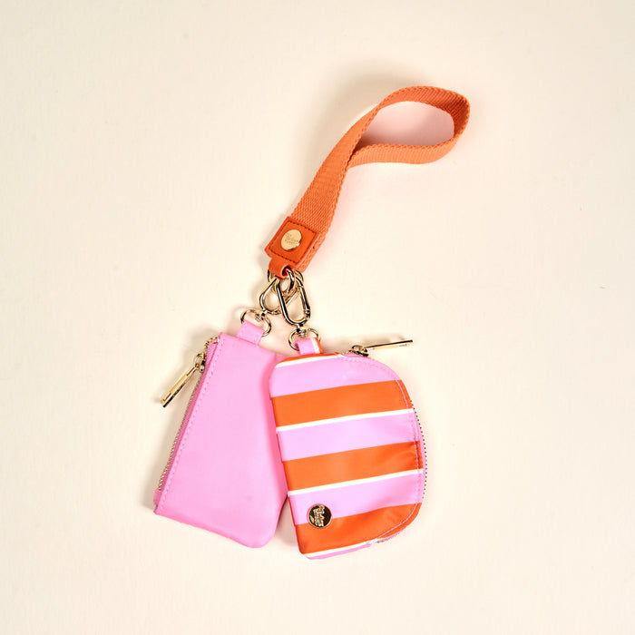 The Darling Effect’s Sicily Stripes Pink Dynamic Duo Pouch Wristlet features two zippered pouches—solid pink and pink with orange and white stripes—joined by metal clasps on an orange strap for a chic, hands-free way to carry essentials.