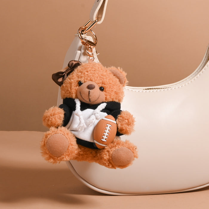 The Darling Effect’s Touchdown Teddy Teddy Bear Bag Charm—a small teddy bear with a brown bow, dressed in a black-and-white outfit and holding a plush football—adds playful style to any bag when attached to the handle of your white handbag, making it a perfect accessory or favorite charm against any beige backdrop.