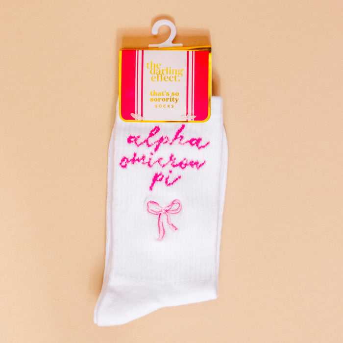 Alpha Omicron Pi Embroidered Crew Socks by The Darling Effect feature pink cursive embroidery and a pink bow on white socks. Packaged in pink, orange, and white—perfect for sorority gifts or embroidered sorority sock lovers.