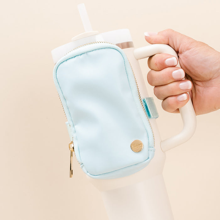 A manicured hand holds a white tumbler with a straw, featuring The Darling Effect's Aqua Tumbler Fanny Pack—a small light blue water-resistant pouch attached to the handle.