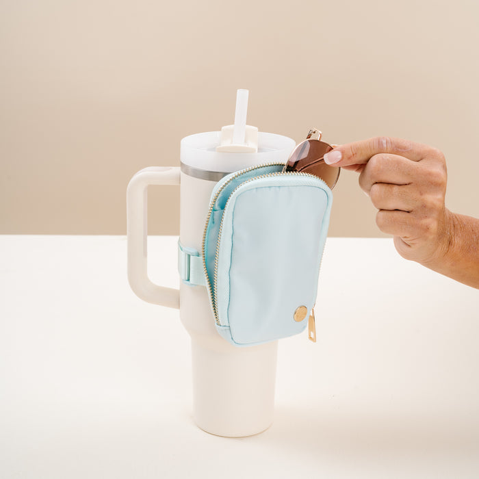 A hand places a key fob into The Darling Effect Aqua Tumbler Fanny Pack in light blue, attached to the handle of a large white tumbler with a straw, set against a neutral background.