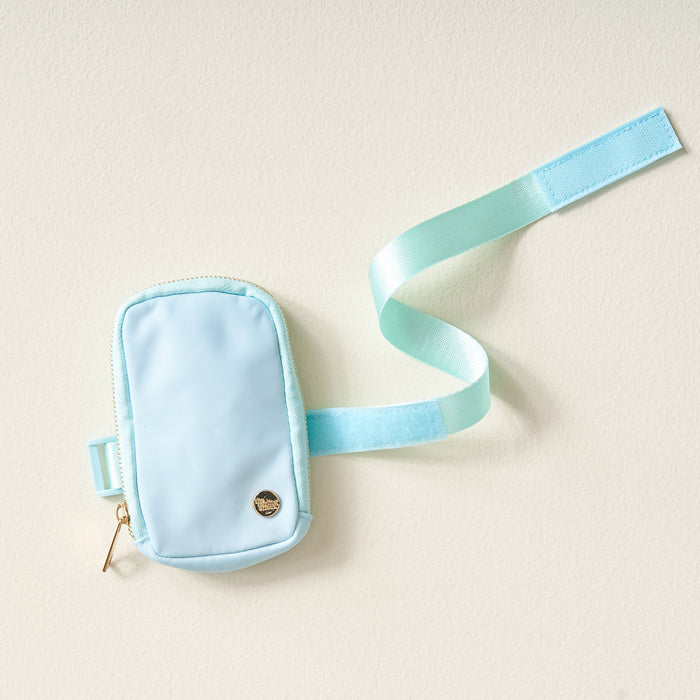 The Darling Effect's Aqua Tumbler Fanny Pack—a small, stylish light blue pouch with a gold zipper and emblem, plus a matching adjustable Velcro strap—rests on a light surface. It's perfect as a water-resistant tumbler fanny pack.