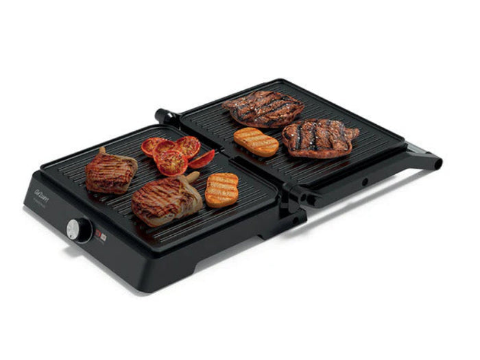 A black dual-grill electric grill shown on a white background, with various foods like steak, tomatoes, and bread cooking on both grates.