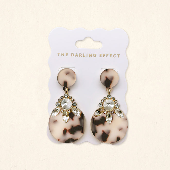 The Darling Effect’s Blonde Tortoise Show Stopper Earrings feature lightweight acetate with circular tops, large round bottoms, gold accents, and clear marquise rhinestones. Presented on a white card branded THE DARLING EFFECT.