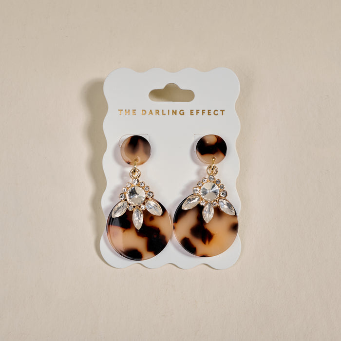 The Darling Effect's Blonde Tortoise Show Stopper Earrings feature rhinestone-embellished acetate in a statement design, displayed on a white THE DARLING EFFECT card with gold text against a beige background.