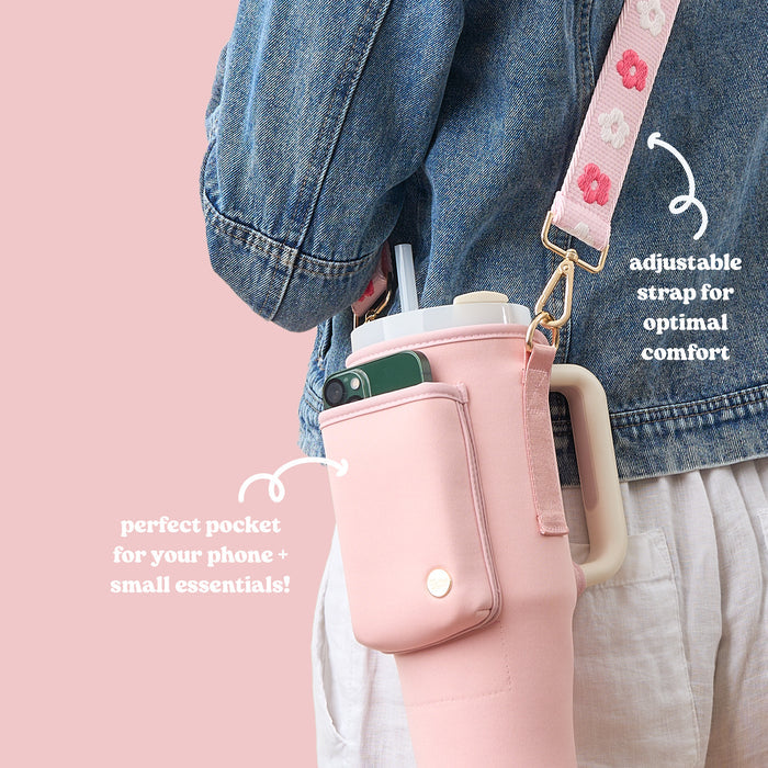 A person in a denim jacket carries The Darling Effect's Blush Tumbler Sling, a pink neoprene bag with an adjustable floral strap and handy pocket for a phone and essentials—combining comfort, style, and functionality.