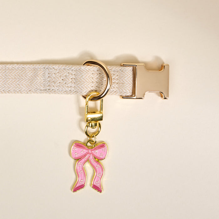 The Darling Effect's Bow Pink Pawfect Collar Charm—a pink bow-shaped charm with gold accents—adorns a beige fabric collar with a gold buckle and D-ring, adding stylish flair to your pet accessory collection.
