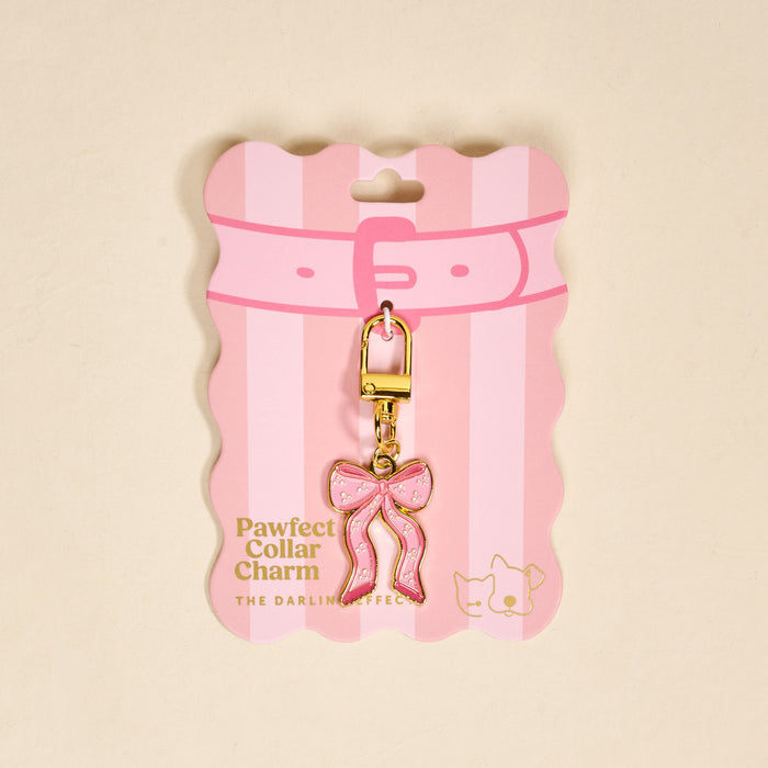 The Darling Effect's Bow Pink Pawfect Collar Charm features a pink bow shape with gold accents and a gold clasp, displayed on pink and white striped packaging—an adorable accessory for any pet collar charm collection.