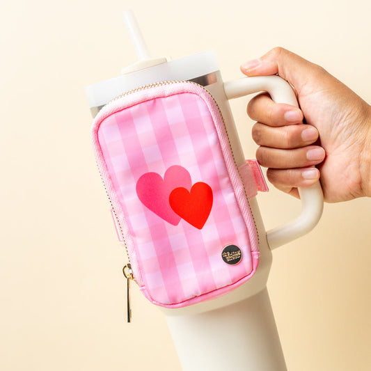 A hand holds a large white tumbler with a handle, accessorized with The Darling Effect's Charming Gingham Tumbler Fanny Pack—a pink checkered pouch featuring two overlapping hearts (pink and red) and a small zipper pull.