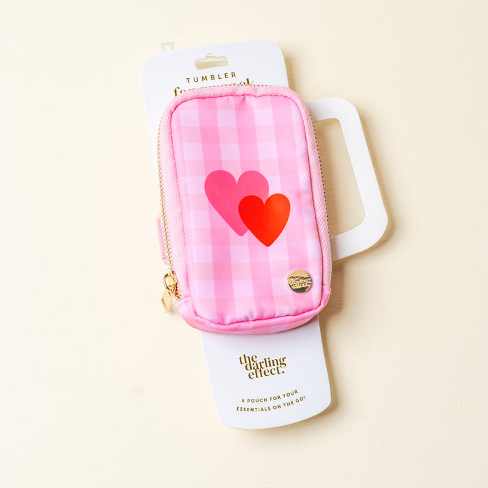 A stylish pink checkered fanny pack, the Charming Gingham Tumbler Fanny Pack by The Darling Effect, features two heart designs, a gold zipper, and a round gold emblem for a chic look, displayed on a pale surface.