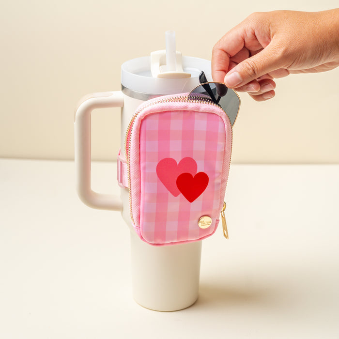 A hand opens The Darling Effect's Charming Gingham Tumbler Fanny Pack in pink plaid with two red hearts, attached to the handle of a white tumbler featuring a straw lid.