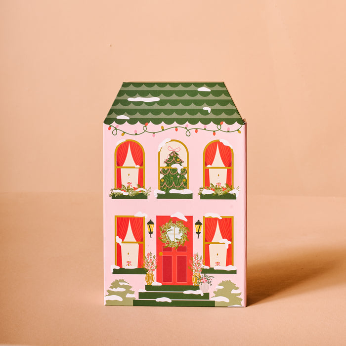 The Darling Effect’s Classic Christmas Ornament Advent Calendar is a decorative box shaped like a festive house—green roof, red door, wreaths, and garlands—perfect for mini ornaments or as a charming accent near your artificial Christmas tree. Set on a plain beige background.