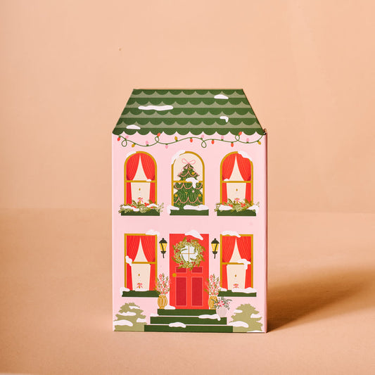 The Darling Effect’s Classic Christmas Ornament Advent Calendar is a decorative box shaped like a festive house—green roof, red door, wreaths, and garlands—perfect for mini ornaments or as a charming accent near your artificial Christmas tree. Set on a plain beige background.