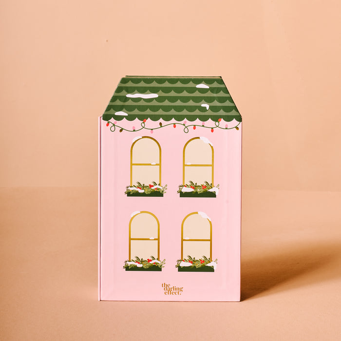 The Darling Effect's Classic Christmas Ornament Advent Calendar features a house-shaped pink box with four yellow-outlined windows, green flower boxes, a scalloped roof with string lights—perfect for displaying mini ornaments. Set against a peach background.