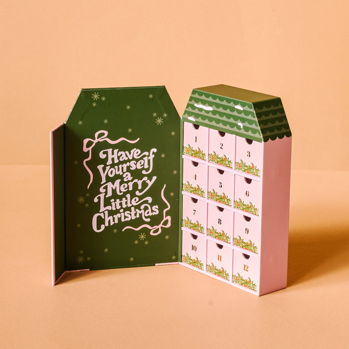 The Darling Effect's Classic Christmas Ornament Advent Calendar is house-shaped with a dark green interior and “Have Yourself a Merry Little Christmas”—ideal for displaying mini ornaments on artificial Christmas trees.