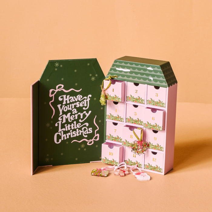 The Darling Effect Classic Christmas Ornament Advent Calendar features numbered drawers, some open to reveal mini ornaments, beside a green door with "Have Yourself a Merry Little Christmas" written on it.