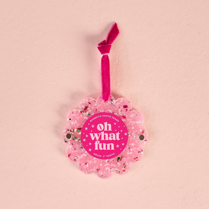 The Darling Effect’s Confetti Pink Scalloped Round Picture Frame Ornament, featuring glitter, a “Oh What Fun” tag, and a pink velvet ribbon, adds playful charm to your decor when hung on a light pink wall.