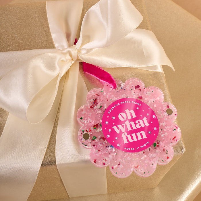 A gold gift box with a white satin ribbon and a pink glittery "oh what fun" tag pairs perfectly with The Darling Effect’s Confetti Pink Scalloped Round Picture Frame Ornament for an extra special touch.
