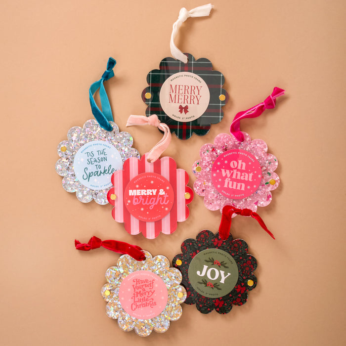 Six holiday gift tags with festive colors, glitter, and messages like "Merry & Bright" are arranged on a tan background—perfect accents for gifts or pairing with The Darling Effect's Confetti Pink Scalloped Round Picture Frame Ornament.