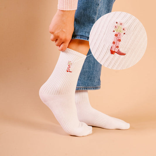 A close-up of The Darling Effect's Cowgirl Boot Embroidered Crew Socks—white crew socks made from breathable fabric, featuring a small pink cowboy boot with flowers, worn with jeans.