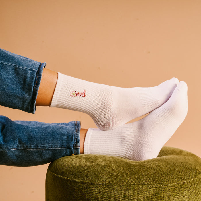 A person wearing The Darling Effect’s Cowgirl Boot Embroidered Crew Socks rests their feet on a green ottoman, dressed in blue jeans against a beige background.