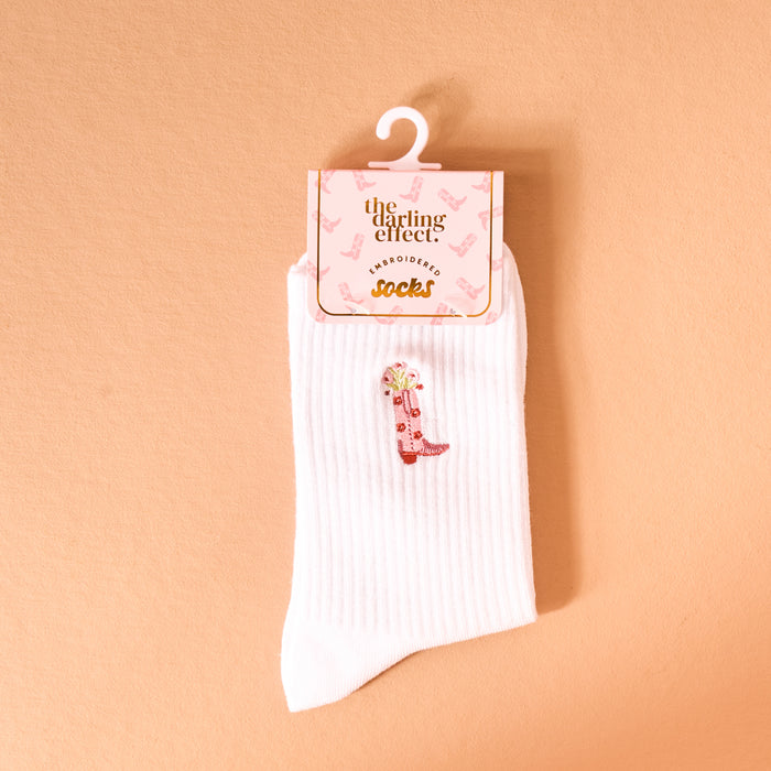 A single white Cowgirl Boot Embroidered Crew Sock from The Darling Effect, featuring a pink boot design, hangs from branded packaging against a peach background. Made with breathable fabric for comfort.