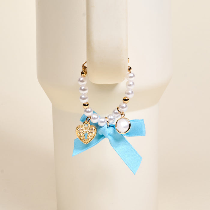 The Darling Effect Daydreamer Tumbler Charm features pearls, gold beads, a heart-shaped lock charm, and a blue ribbon bow, making it an adorable accessory for your favorite drinkware.