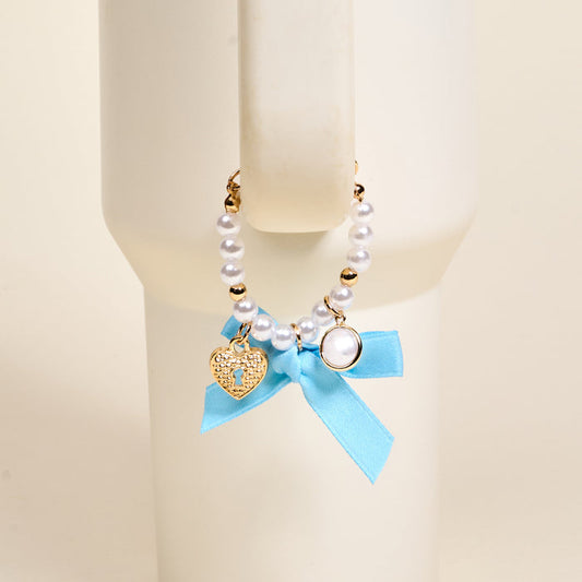 The Darling Effect Daydreamer Tumbler Charm features pearls, gold beads, a heart-shaped lock charm, and a blue ribbon bow, making it an adorable accessory for your favorite drinkware.