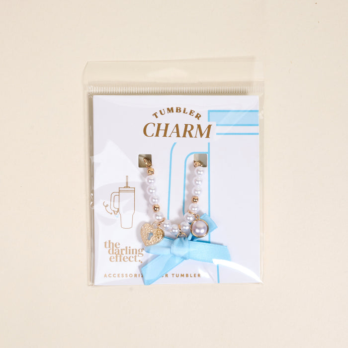 The Darling Effect's Daydreamer Tumbler Charm features white and gold beads, a gold heart, pearl, blue ribbon, and comes in a clear bag with a card labeled “Tumbler Charm” and “The Darling Effect.”.