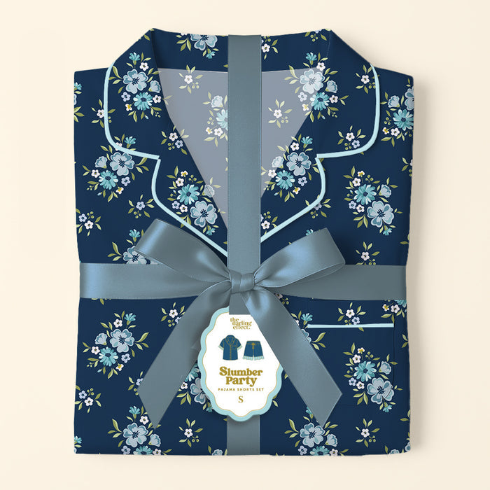 The Darling Effect’s Endless Daydream Navy Slumber Party Pajama Shorts Set features folded, ultra-soft navy pajamas with a light blue floral pattern, tied with a satin ribbon and bow. Gift-ready, it includes a tag with an illustration of the set.