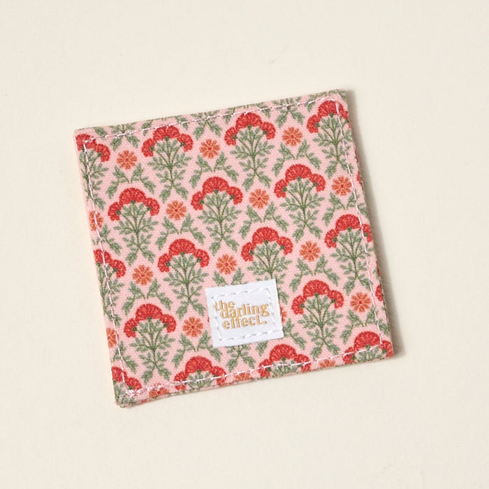 The Darling Effect’s Everbloom Dusty Rose Corner Keeper Fabric Bookmark features a red and green floral pattern on a light background with a gold logo label—perfect as a unique book lover gift or paired with a matching coaster.