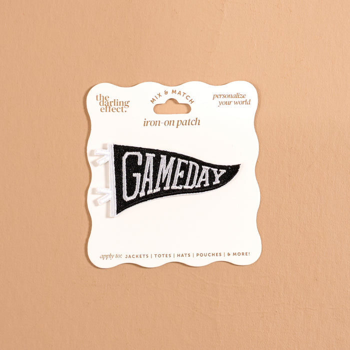 The Darling Effect Gameday Black Mix & Match Iron-On Patch is a black and white pennant-shaped patch featuring the word "GAMEDAY," displayed on card packaging against a beige background—ideal for customizing your favorite items with this unique, stylish iron-on accent.
