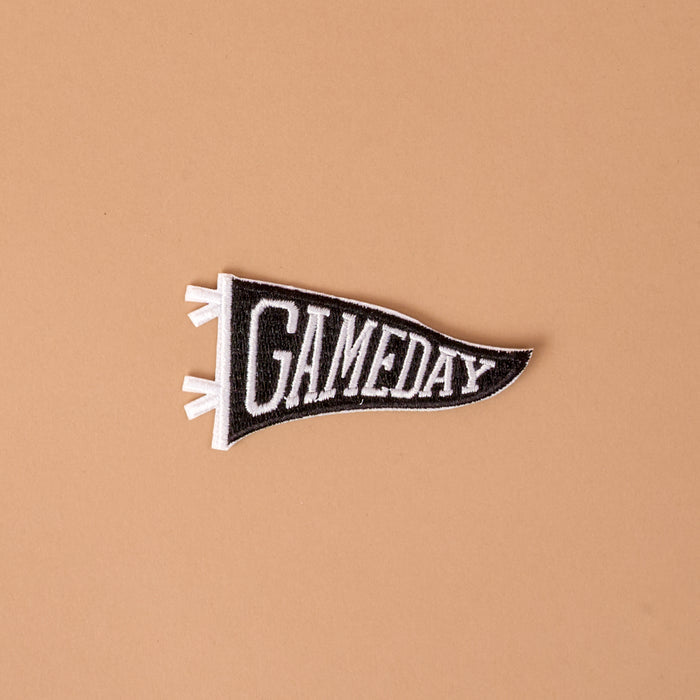 Introducing the Gameday Black Mix & Match Iron-On Patch by The Darling Effect—a black and white felt pennant patch featuring the word "GAMEDAY" in bold capital letters on a simple beige background, perfect for customizing your favorite items with a unique and stylish touch.