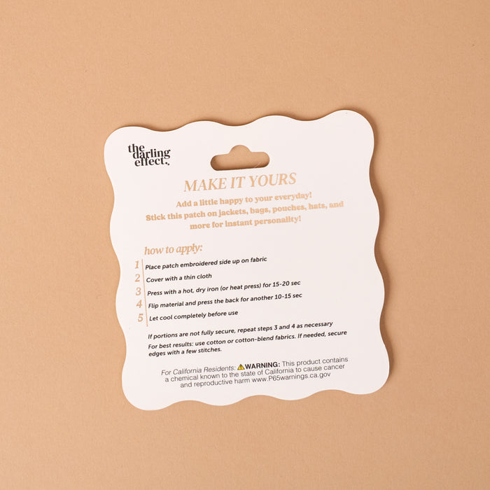 A product instruction card titled "MAKE IT YOURS" from The Darling Effect explains how to apply the Gameday Black Mix & Match Iron-On Patch using a hot iron, featuring colorful designs, safety tips, and a warning for California residents at the bottom. The instructions are presented on a tan background—making it easy to customize your favorite items.
