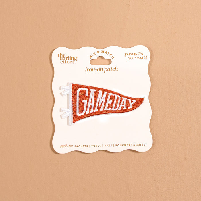 A pennant-shaped iron-on patch from The Darling Effect, featuring "GAMEDAY" in white letters on an orange background, perfect for customizing your look. The Gameday Orange Mix & Match Iron-On Patch is displayed on cream packaging against a tan background.