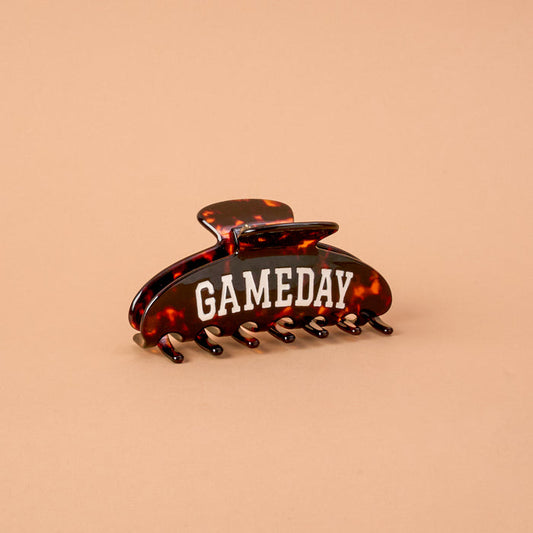 The Gameday Word Up Claw Clip by The Darling Effect, featuring a brown tortoiseshell design with "GAMEDAY" in bold white letters, is displayed on a plain beige background, highlighting its secure hold and statement style.