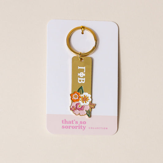 The Darling Effect's Gamma Phi Beta Enamel Keychain, featuring Greek letters and colorful enamel flowers, is displayed on a white card labeled "that's so sorority collection" in pink—perfect for any sorority gift.
