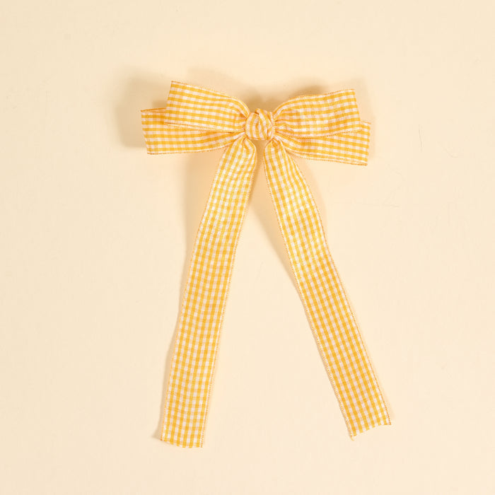 The Darling Effect's Gingham Yellow Double Take Hair Bow features yellow and white gingham ribbon tied into a bow with two long tails, set against a light beige background—perfect for hair accessories or as a stylish bow clip.