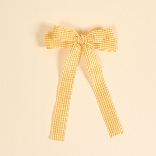 The Darling Effect's Gingham Yellow Double Take Hair Bow features yellow and white gingham ribbon tied into a bow with two long tails, set against a light beige background—perfect for hair accessories or as a stylish bow clip.