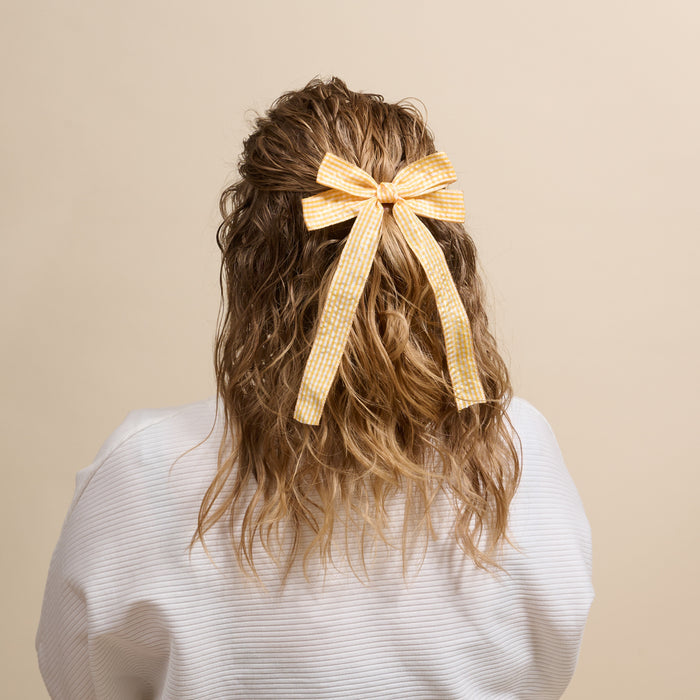 A person with wavy, shoulder-length hair wears The Darling Effect’s Gingham Yellow Double Take Hair Bow over a white shirt. Featured from behind against a plain beige background, the bow is a stylish, standout accessory.