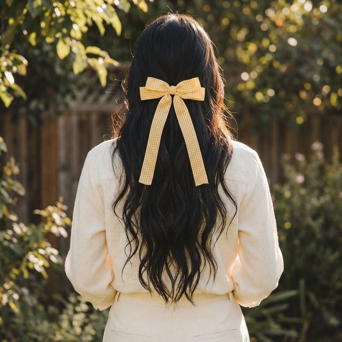 A person with long, wavy dark hair stands outdoors in cream, wearing The Darling Effect's Gingham Yellow Double Take Hair Bow, its cheerful pattern highlighted by sunlight against the greenery and a wooden fence.