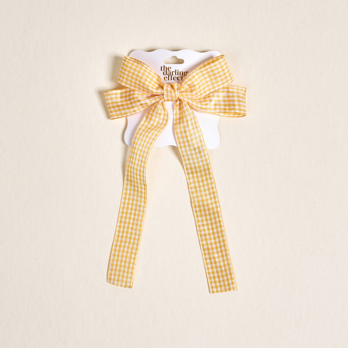 The Darling Effect’s Gingham Yellow Double Take Hair Bow, featuring yellow and white gingham long tails, is displayed on a scalloped card—ideal for lovers of charming hair accessories.