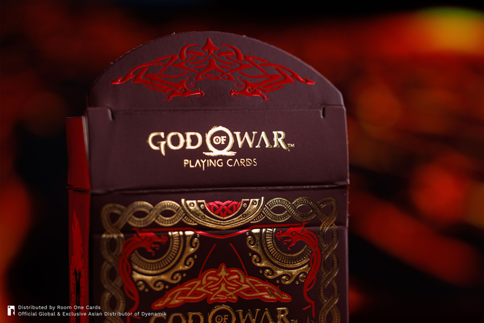 God of War Playing Cards box in dark purple with red and gold ornate designs, shown against a blurred warm background.