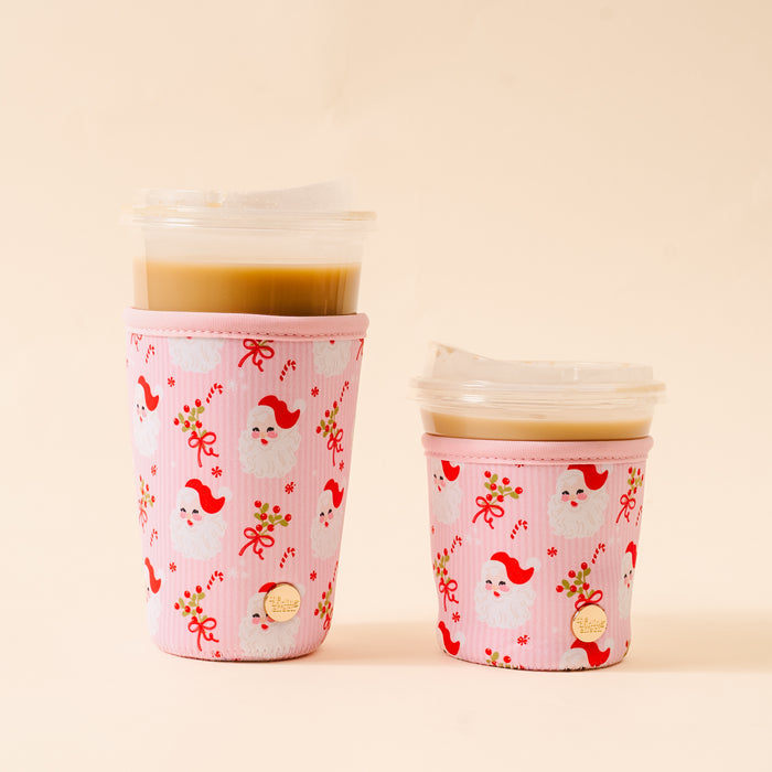Two takeaway iced coffees with clear lids, each wrapped in The Darling Effect's Holly Jolly Santa Sip Savvy Drink Sleeve featuring Santa faces, holly, and red bows, set against a plain light beige background.