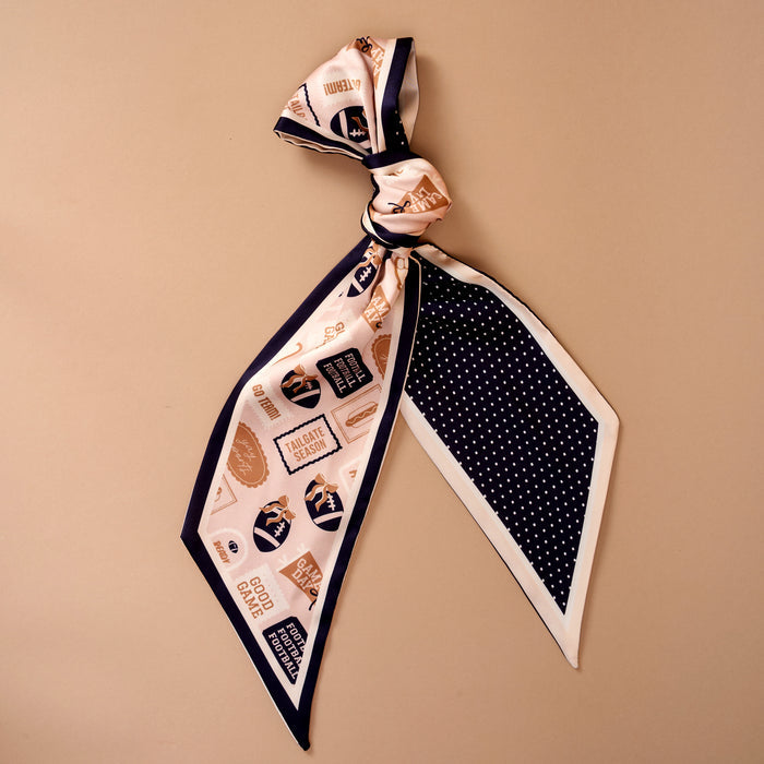 Introducing the Huddle Up Twirl & Tie Scarf Accessory from The Darling Effect—a sophisticated satin scarf accentuated by a stylish knot at the top. This accessory showcases printed labels and graphics in elegant navy, white, and gold. One end features crisp white polka dots on a navy background, while the other displays assorted motifs set against cream, offering endless options for versatile styling.