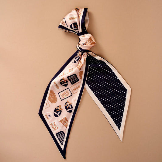 Introducing the Huddle Up Twirl & Tie Scarf Accessory from The Darling Effect—a sophisticated satin scarf accentuated by a stylish knot at the top. This accessory showcases printed labels and graphics in elegant navy, white, and gold. One end features crisp white polka dots on a navy background, while the other displays assorted motifs set against cream, offering endless options for versatile styling.